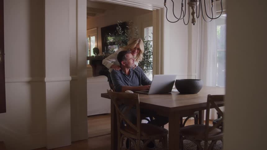 A woman walks in and hugs a man from behind as he sits at the dining table working on his laptop at midday