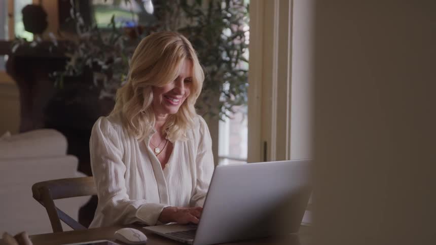 Smiling blonde woman works on a laptop at a wooden table