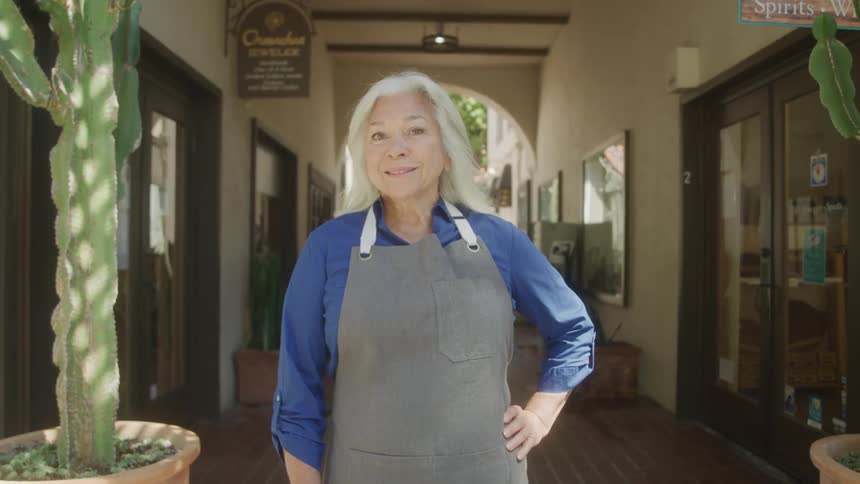 The video shows an elderly woman with gray hair and an apron standing in the doorway of a shop she has a warm smile and is looking directly at the camera