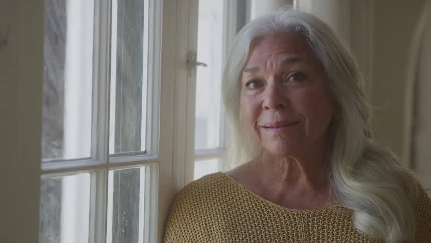 A woman with long gray hair stands peacefully by a sunlit window in a mustard sweater