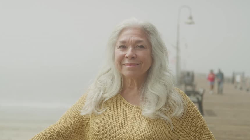 The video shows a closeup of an elderly woman with long gray hair and a yellow sweater she is standing outside on a foggy day smiling happily at the camera