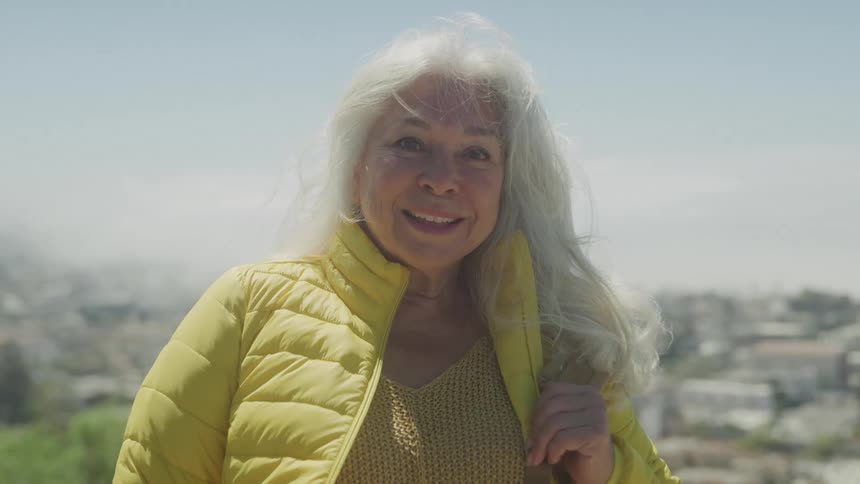An elderly woman with gray hair wearing a yellow winter jacket smiles warmly while standing outdoors on the outskirts of the city at midday