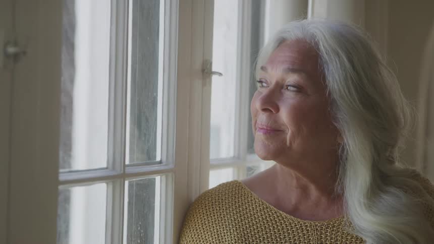 The video shows a closeup of an elderly womans face with long gray hair she is looking out a window with a calm and peaceful expression on her face