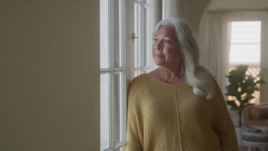 The video shows a closeup of an elderly woman with long gray hair looking out a window she has a calm and peaceful expression on her face