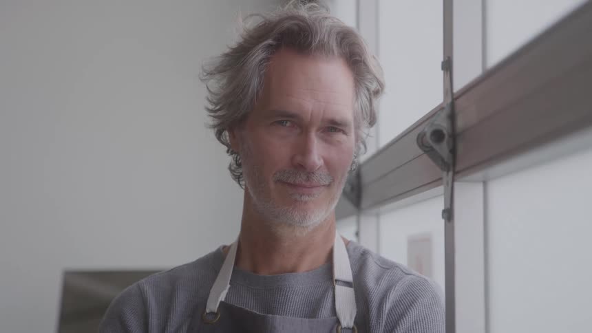A man with saltandpepper hair and an apron smiles gently while standing by a large window