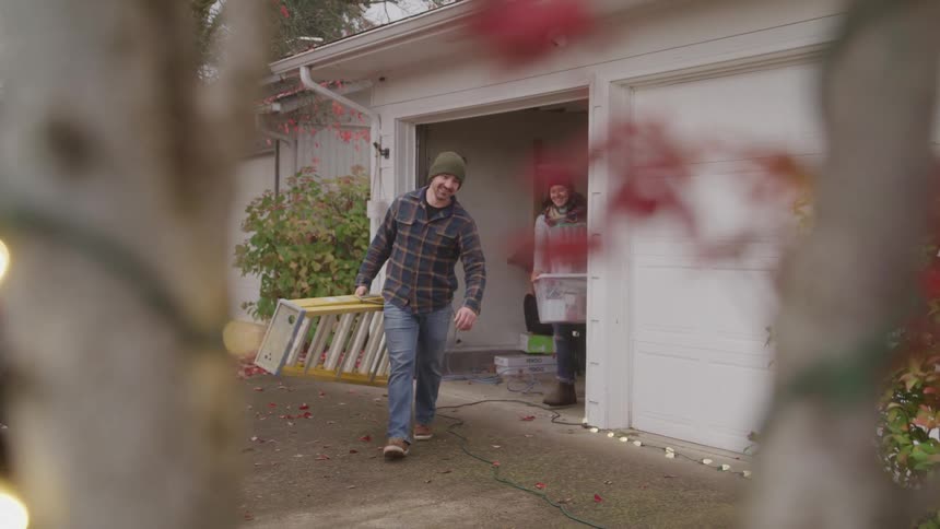 A man walks out of a garage holding a ladder followed by a woman carrying two large boxes as they both smile