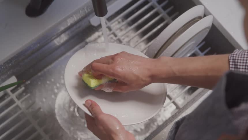 A person washes a plate in the kitchen sink with a sponge at midday
