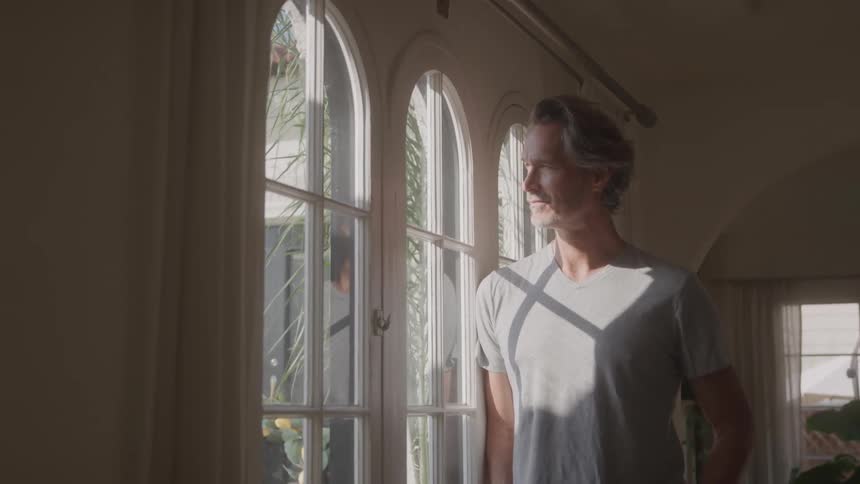 A middleaged man stands beside a glass window staring outside the window at midday