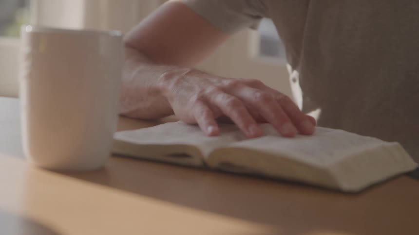 A person wearing a grey shirt sits at a table and flips the pages of an open book next to a white mug