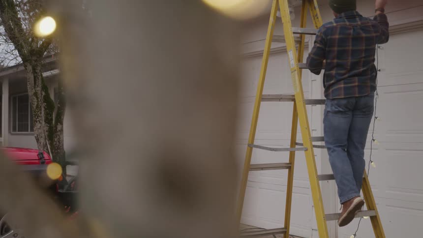 A man climbs a ladder outdoors in front of his garage hanging christmas lights on his roof at sunset while his wife points to the roof directing him