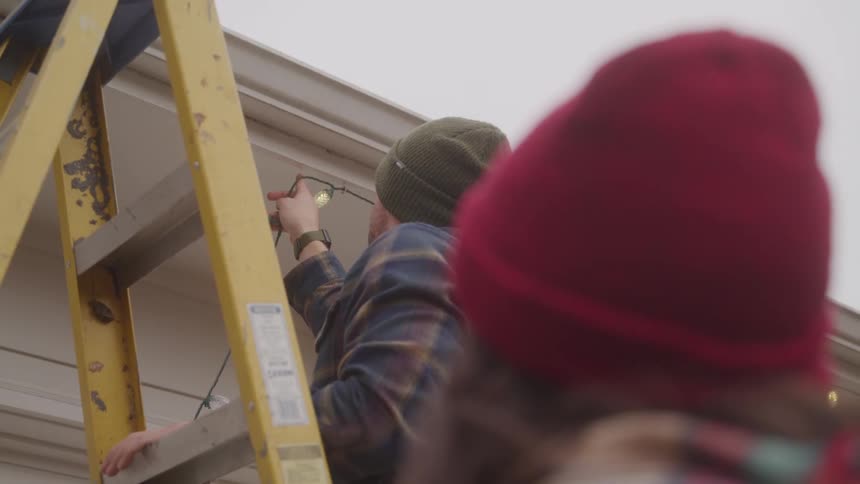 A man climbs up a ladder to fix christmas lights on the roof while a woman stands nearby watching him at midday