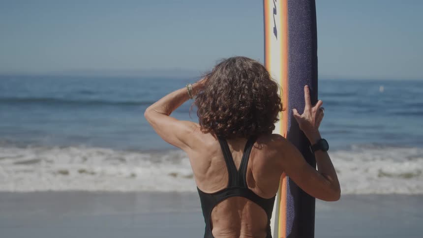 A woman looks out over the water while holding her surfboard