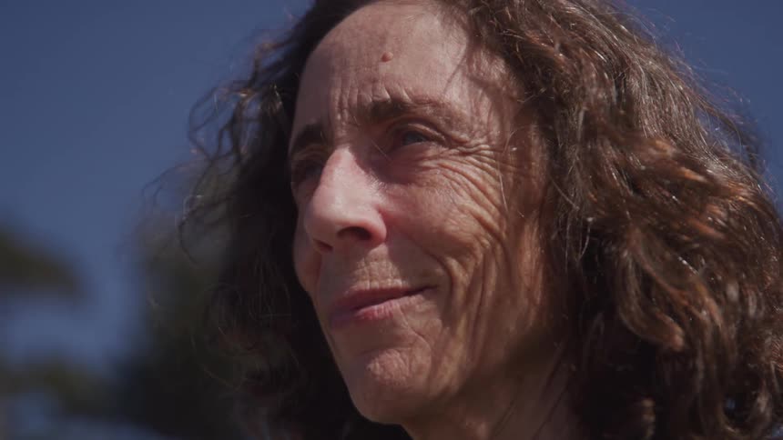 An elderly woman with long hair stands outdoors with a smile on her face