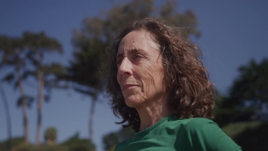 An elderly woman with long hair stands outdoors with a bit of sadness on her face
