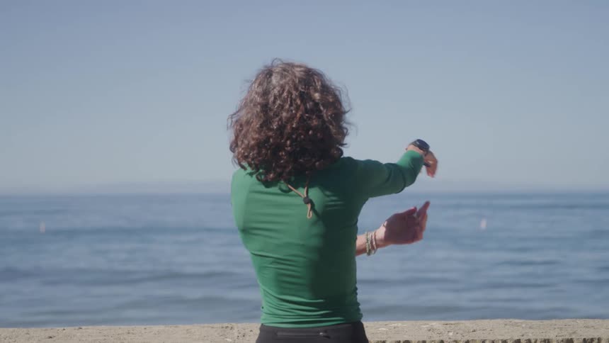 A woman with long curly brown hair wears a green long sleeved shirt and black leggings while stretching her arms in front of a calm ocean