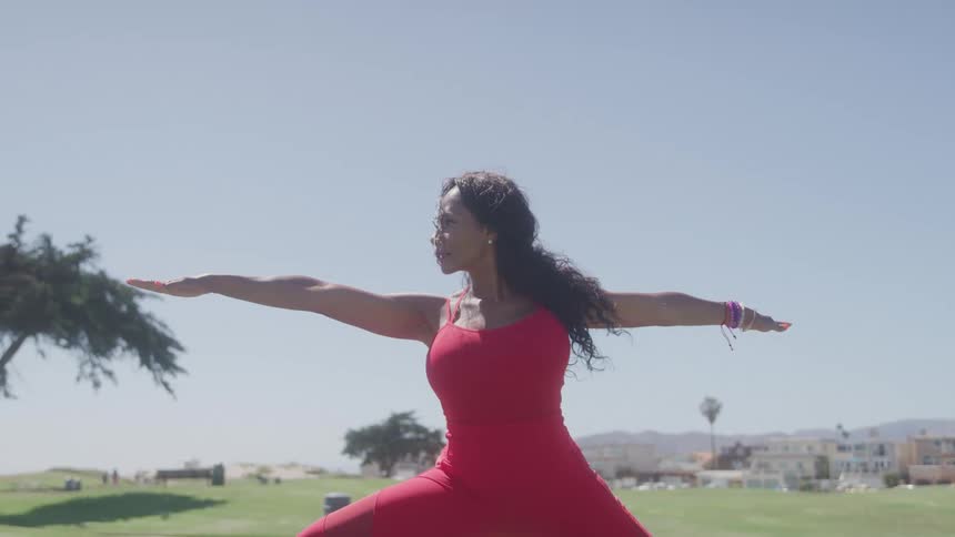 The video shows a woman performing a yoga pose in a park she is wearing a red outfit and has her arms extended in a warrior pose
