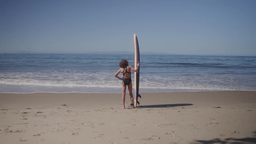 A person holding a surfboard stands on the beach at midday watching the ocean waves