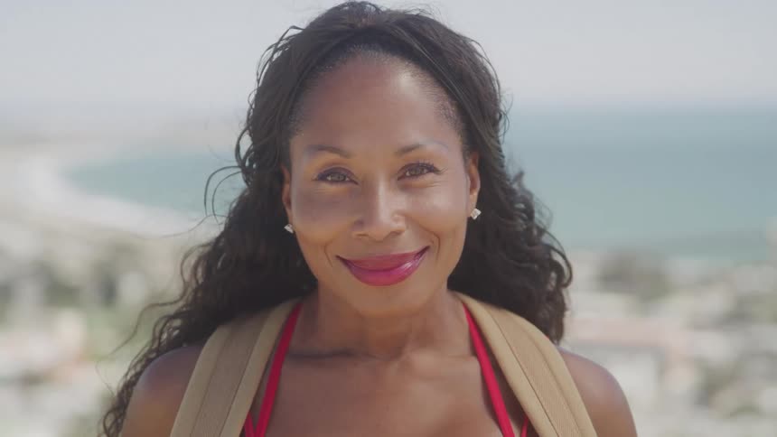 A middleaged woman stands outdoors at midday with a smile on her face wearing red lipstick