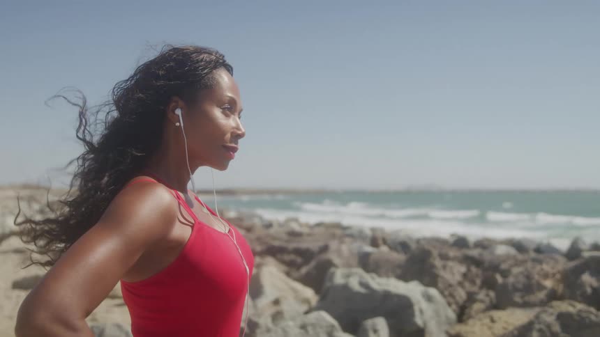 A fit woman in a red top listens to music through earphones while standing on a rocky beach her hair blowing in the wind as she looks out at the ocean