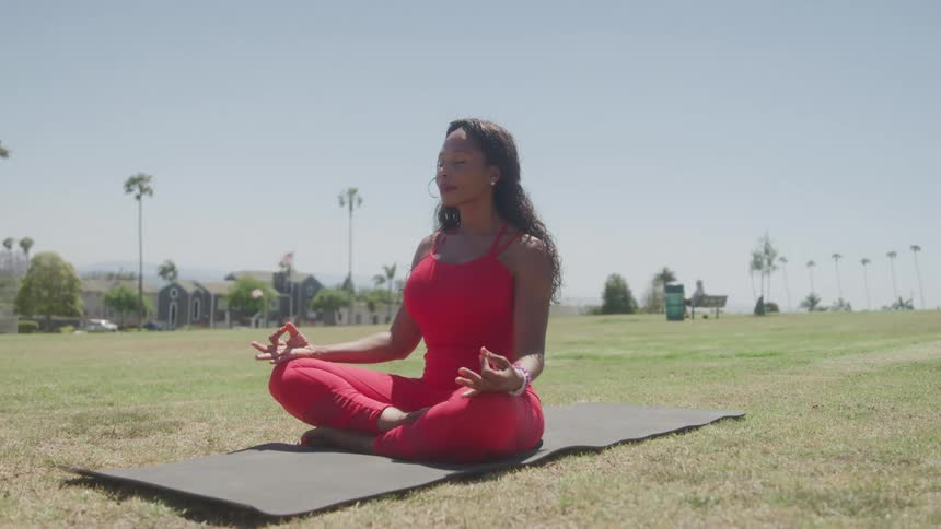 Woman in a red jumpsuit practices meditation sitting on a mat on the grass
