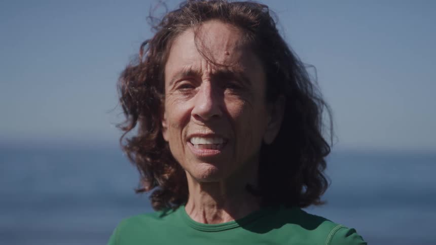 A older woman with white hair and a green shirt is smiling and looking at the camera on a windy day with the ocean and sky in the background