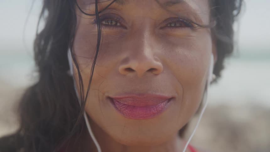 A woman with long dark hair and earbuds is looking directly at the camera with a gentle smile her face is illuminated by bright sunlight and water droplets are visible on her hair and face