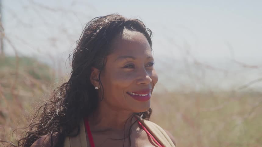 A woman smiles warmly in the sunlight with wind blowing through her hair in a dry open field