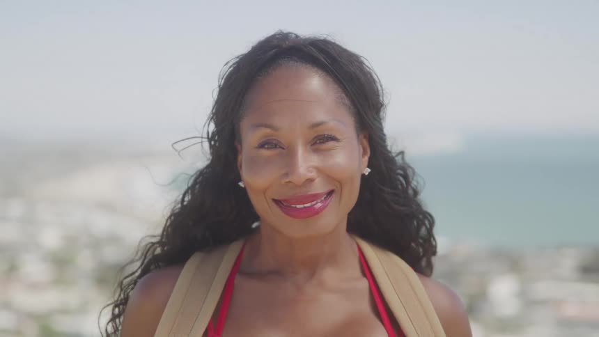The video shows a smiling woman with long dark hair wearing a red bikini top and a beige coverup she is looking directly at the camera with a happy expression and a body of water is visible in the background