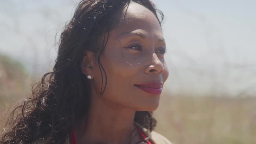 The video shows a closeup of a woman with long dark curly hair smiling as she looks offscreen she is standing in a sunny outdoor setting with some foliage in the background
