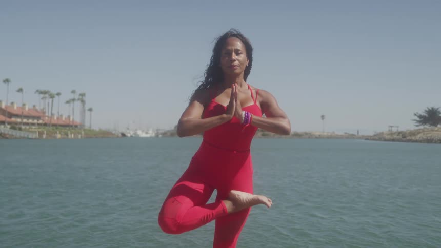 The video shows a woman performing a yoga pose near a body of water she is wearing a red outfit and has her arms raised to her chest