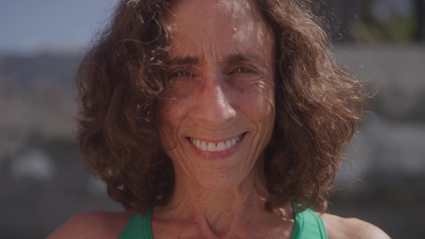 A joyful older woman with curly brown hair looks directly into the camera with a wide genuine smile on her face