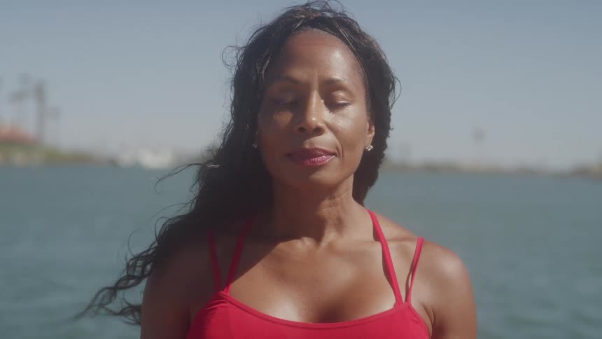 A serene woman in a red top stands with her eyes closed her hair blowing in the wind with a body of water in the background