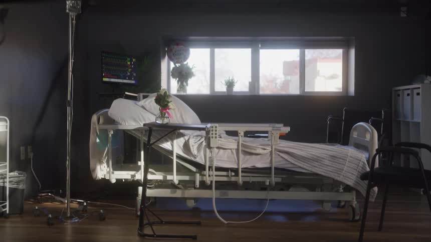 A sunlit hospital room holds an empty bed surrounded by medical equipment flowers and quiet stillness