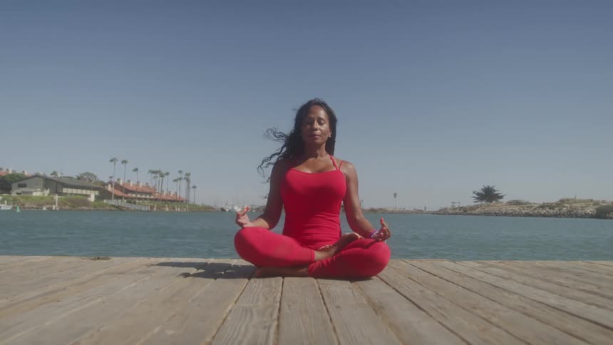 A young black woman performs yoga on a wooden raft outdoors during daytime