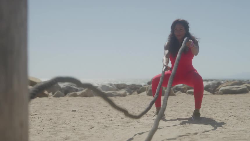 A woman in a red jumpsuit performs a workout with heavy ropes on a sandy beach with rocks and the ocean in the background under a clear sky