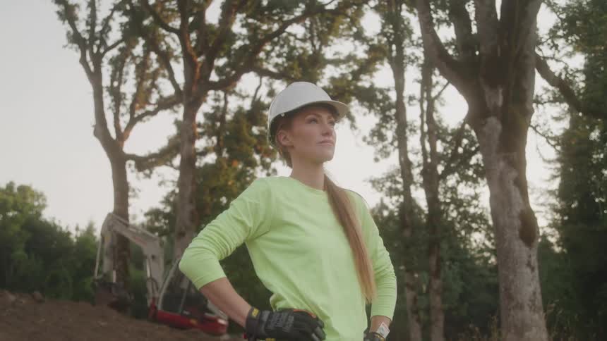 The video shows a woman wearing a hard hat and gloves standing in a clearing with her hands on her hips she is looking at the ground and a construction vehicle is visible behind her