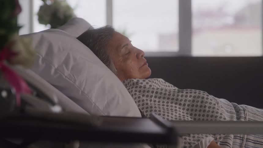 An elderly person with gray hair is lying in a hospital bed looking up with a neutral expression the person is wearing a patterned hospital gown and a pillow is propped behind their head
