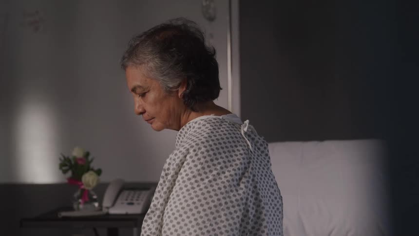 An elderly female patient sits contemplatively at the edge of a bed in a hospital room