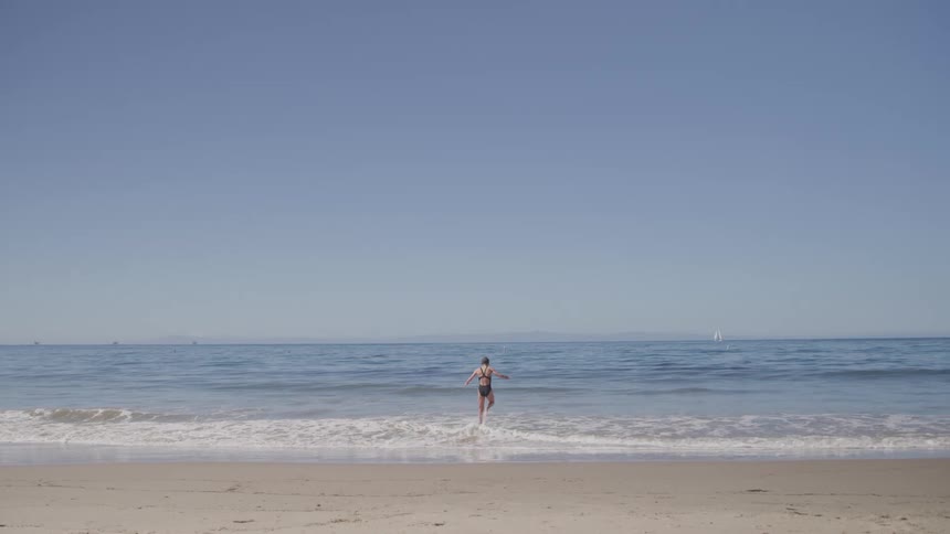 The video shows a wide shot of a person standing on a sandy beach facing the ocean they are wearing a dark bathing suit and looking out at the waves crashing on the shore