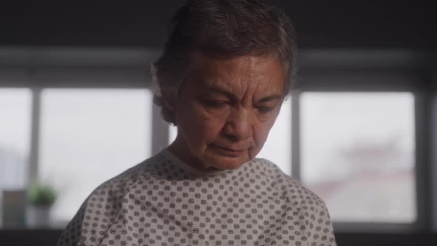 An elderly female patient in a hospital gown looks downward in a softly lit room