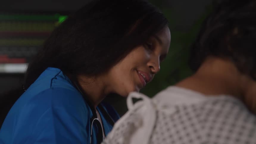 A medical professional in blue scrubs smiles and speaks to a patient who is wearing a hospital gown and has their back to the camera