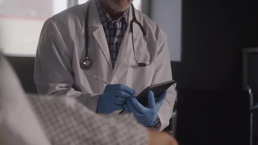 Doctor with gloves taking notes on a tablet and consulting a patient lying down in a hospital