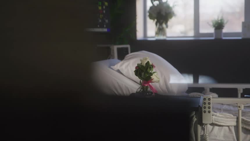 A vase with white flowers sits on a table next to an empty hospital bed with a remote control hanging from the side of the bed