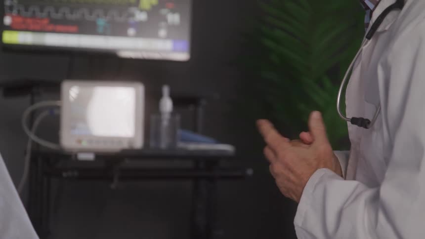 Doctor in a white coat gestures with his hands in a room with medical equipment