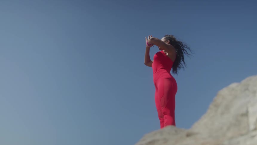 A woman in a form fitting red top and leggings stands on a rock on a sunny day and raises her hands over her head before bringing them down to a prayer position