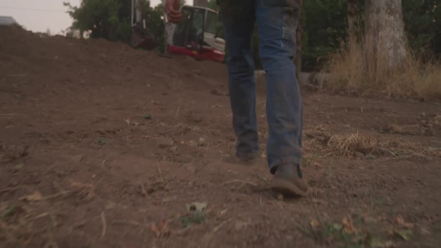 A man in jeans and work boots walks away on a dirt path surrounded by grass and construction equipment
