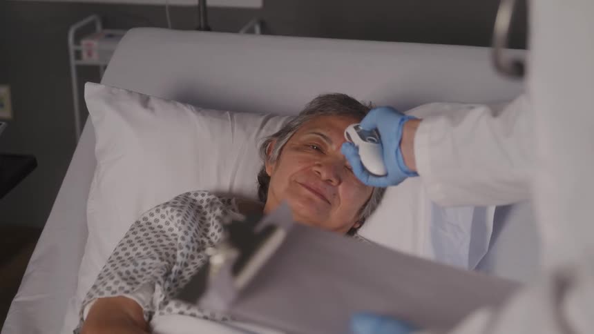 A doctor checks the temperature of a smiling elderly woman resting in a hospital bed