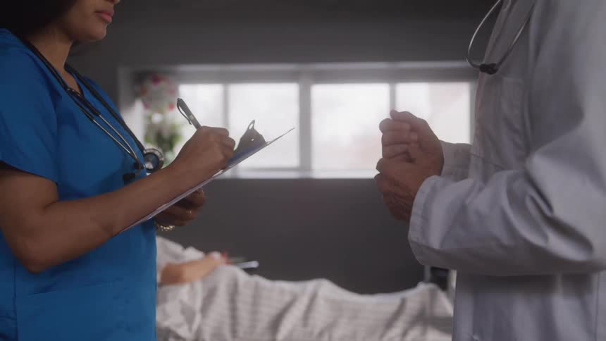 A female medical professional takes notes from another male doctor standing with her in a hospital room during daytime