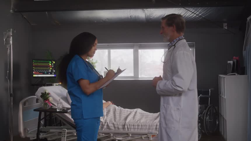The video shows a male and female doctor discussing a case in a hospital room a patient is visible in the background lying in a bed