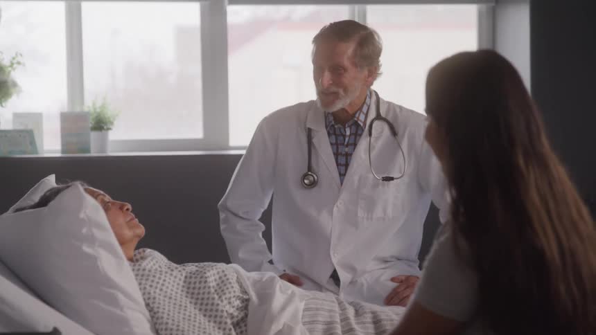 Doctor talks with patient in hospital bed as visitor sits nearby